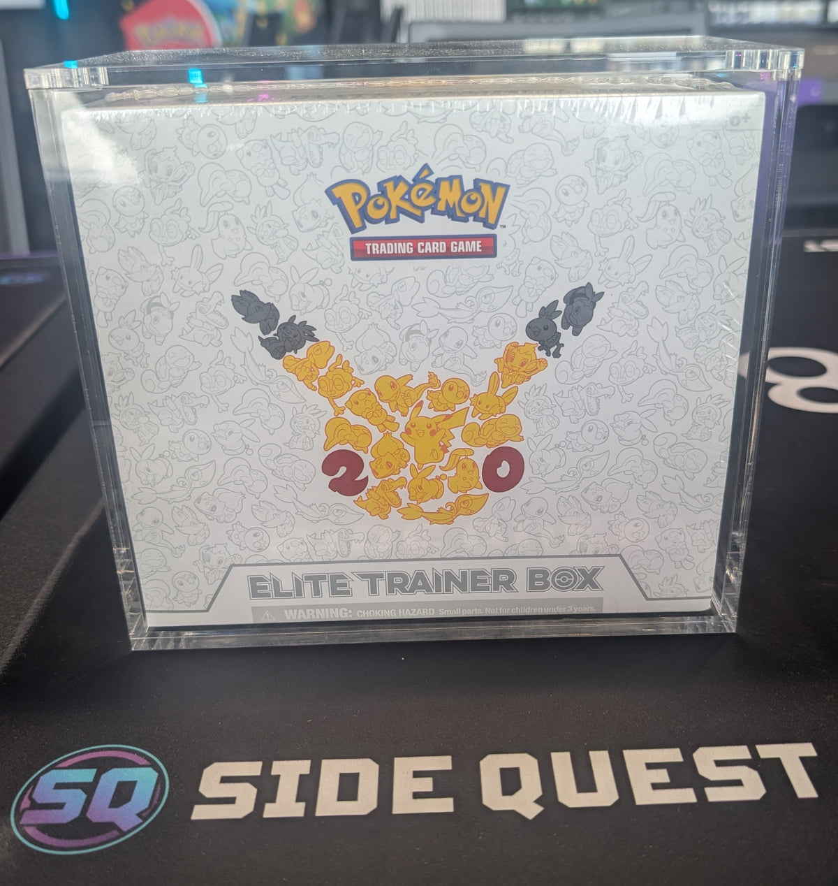 A sealed Pokémon Elite Trainer Box displayed in a clear plastic case on a black desk mat with the Side Quest logo, with a Pokémon store interior blurred in the background.